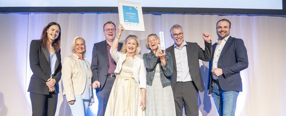 Gruppenbild mit sieben Personen. Dame in der Mitte hält Urkunde in die Höhe.
