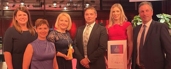 6 Personen stehen in Abendkleidung nebeneinander. Die zweite von rechts hält eine gerahmte Urkunde.