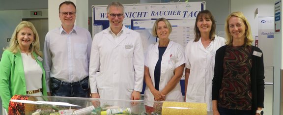 6 Personen stehen hinter Vitrine in Form einer liegenden Zigarette