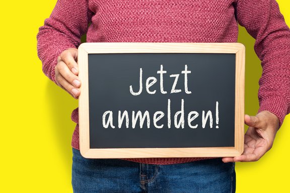 schwarze Tafel mit Kreideschrift "jetzt anmelden"", gehalten von einem Mann mit Jeans und rotem Pulli
